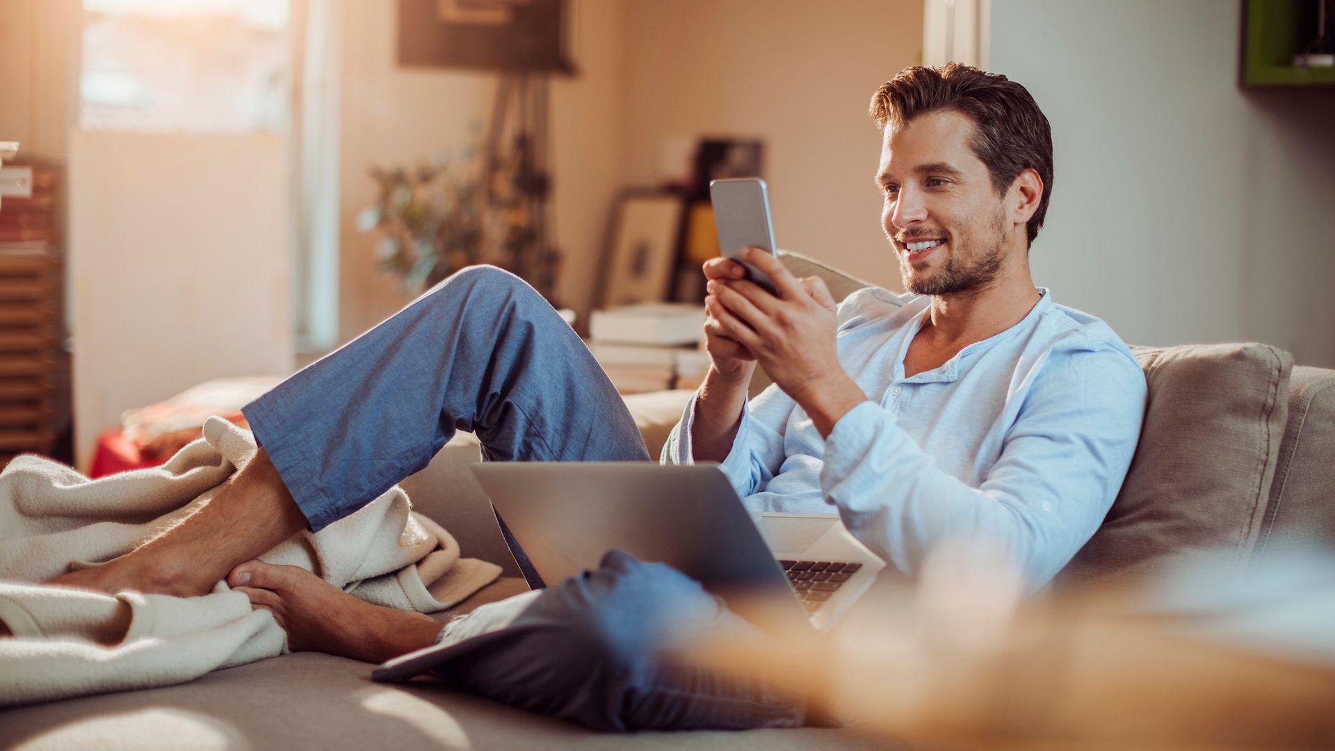 Ein junger Mann sitzt auf dem Sofa mit Handy und Laptop